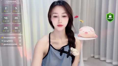 Media: Video of an East Asian woman with long brown hair in a braid, wearing a sheer blue nightgown with black trim, standing against white curtains.