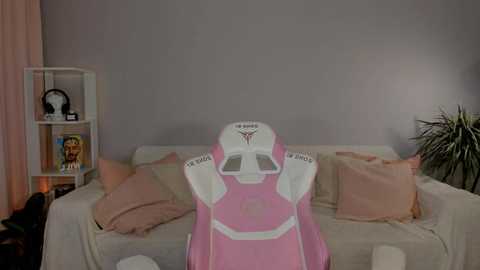 Media: Video of a pink, white, and gray gaming chair with \"Aerofly\" logo, positioned on a cream sofa with soft cushions, in a room with a white wall, green plant, and a white shelving unit with books and toys.