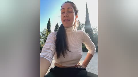 Media: Video of a smiling Latina woman with long black hair, wearing a white ribbed turtleneck and black pants, standing outdoors in front of tall trees on a sunny day.