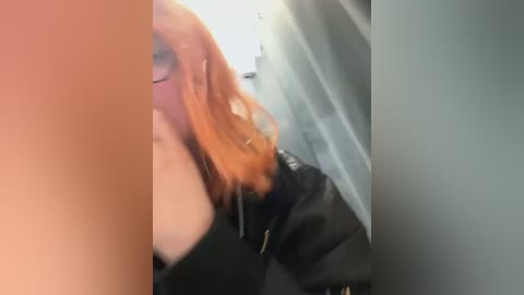 Media: A video of a woman with long, straight, orange hair, wearing a black jacket, partially obscured by a large, blurry object. She has a fair complexion and is smiling.