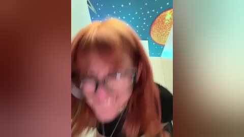 Media: Video of a young woman with reddish-brown hair, wearing glasses and a black shirt, looking directly at the camera with a slight smile, in a room with a blue starry wallpaper and a sun-shaped poster.