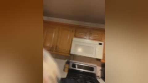 Media: Blurry video of a kitchen with light-colored wooden cabinets, white microwave, and black stove, partially obscured by a person in the foreground.