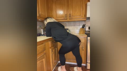 Media: Video of a blonde woman in black leggings and a hoodie, bending over in a wooden kitchen, showcasing her curvy figure and buttocks.