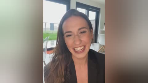 Media: Video of a smiling woman with light skin and long brown hair, wearing a black jacket, indoors near a window with a green garden outside.