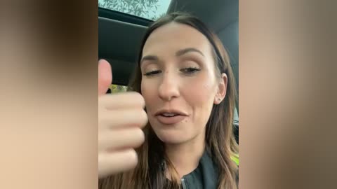 Media: Video of a fair-skinned woman with brown hair, wearing a black top, giving a thumbs-up gesture from inside a car. The background features a blurry view of trees outside.