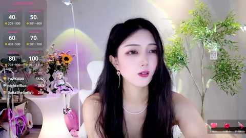 Media: Video of an East Asian woman with long black hair, wearing a white dress and pearl necklace, sitting in a modern room with greenery and floral arrangements.