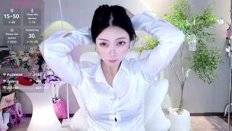 Media: A video of an East Asian woman with fair skin, straight black hair, and light makeup, sitting in a white chair, adjusting her hair. The background features a wall with posters and a potted plant.