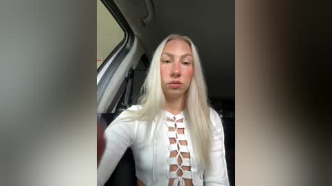 Media: Video of a fair-skinned woman with long, straight platinum blonde hair, wearing a white, long-sleeve, lace-up top, standing in an SUV's open door.