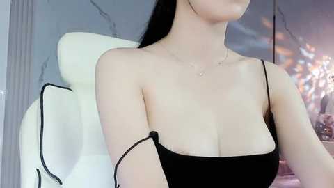 Media: Video of a fair-skinned, slender woman with long black hair, wearing a black spaghetti-strap top revealing her cleavage, sitting in a modern, white armchair. Background features a gray, textured wall with soft, warm lighting.