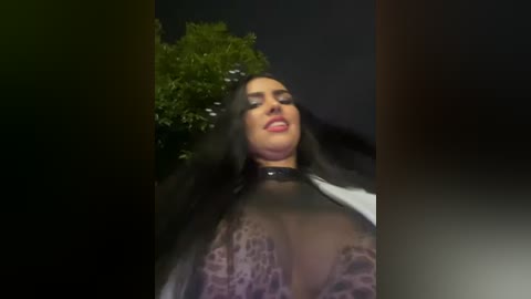 Media: A video of a woman with long black hair, wearing a sheer black lace top, standing in front of a dark background with a leafy tree visible. Her makeup is bold, with red lipstick and dramatic eye makeup.