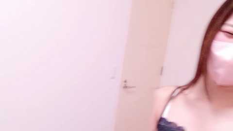 Media: A video of a partially visible young woman with fair skin, wearing a black lace bra and a white mask, standing in a dimly lit, minimalist room with a white door.