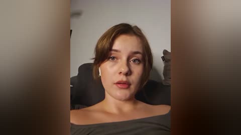 Media: Video of a young Caucasian woman with short brown hair, wearing an off-shoulder gray top, sitting indoors, blurred background, neutral expression.