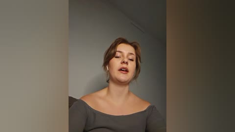 Media: Video of a young Caucasian woman with light brown hair, wearing a grey off-shoulder top, singing or speaking with eyes closed, standing in a dimly lit room with plain walls.