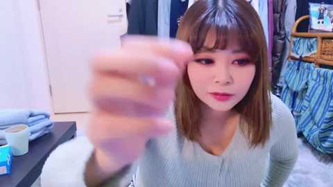 Media: Video of a young East Asian woman with straight brown hair, light skin, and red lipstick, wearing a light green sweater, standing in a modern bedroom with blue-striped sheets, a wicker chair, and a white door.