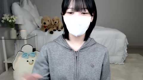 Media: Video of an Asian woman with short black hair and a white mask, wearing a gray hoodie, holding a white teddy bear with blue ribbon. Background features a bed with plush toys and a vase of flowers.