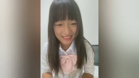 Media: Video of an Asian girl with long black hair, wearing a white blouse with a pink bow, smiling broadly with eyes closed. Background is a plain, light-colored wall.