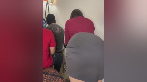 Media: Video of three individuals, two men and one woman, sitting in a small, dimly lit room. The woman, with a curvy figure, wears a tight gray skirt and a maroon top.