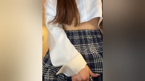 Media: A video of a woman's torso and thighs, wearing a white cropped sweater and a plaid skirt, standing against a beige wall.