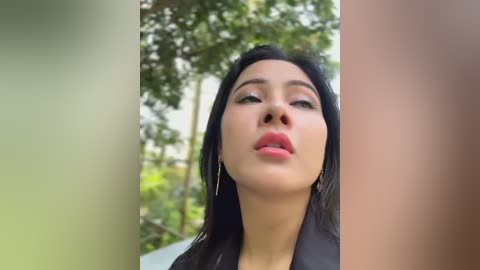 Media: Video of a young woman with long black hair, fair skin, and full lips, wearing a black jacket, against a blurred green background with trees.