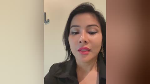 Media: Video of a woman with medium skin tone, long black hair, and wearing a black jacket, standing in a doorway with a beige wall and brown door on the right. She has bold makeup and a confident expression.