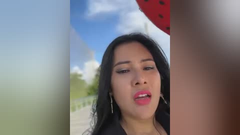 Media: A video of a Latina woman with long black hair, wearing a black top and hoop earrings, speaking outdoors against a blurred background of greenery and a red watermelon slice.