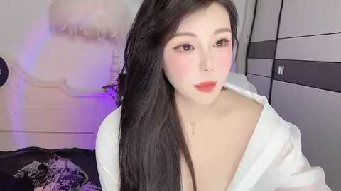 Media: Video of a young Asian woman with long, straight black hair, wearing an unbuttoned white shirt, red lipstick, and subtle makeup. She stands in a modern bedroom with a white wall and a black and white dresser.