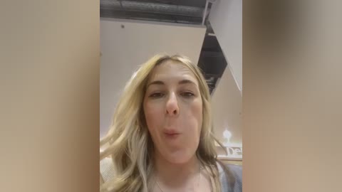 Media: A video of a blonde woman with a pouty face, wearing a gray top, in a modern office with beige walls, metal ceiling, and white furniture.