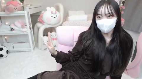 Media: Video of a young Asian woman with long black hair, wearing a white face mask, brown jacket, and brown pants, seated in a pastel pink room with plush toys, shelves, and a white chair.