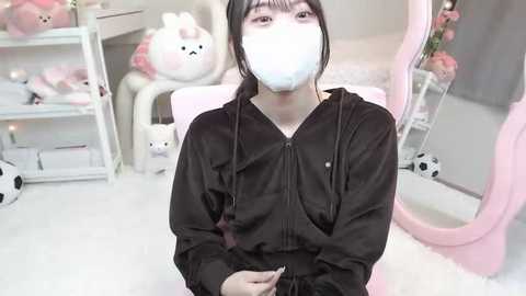 Media: A video of an East Asian woman with a white surgical mask, black hair in a ponytail, wearing a black hoodie, seated in a pink gaming chair with a plush bear behind her.