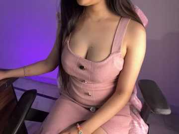 Media: Video of a light-skinned woman with long, straight black hair, wearing a pink denim overall dress, sitting on a black office chair against a purple-lit background.