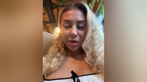 Media: A video of a light-skinned woman with long, wavy blonde hair, wearing a white off-shoulder top with black trim. She has a serene expression, eyes closed, in a dimly lit indoor setting with greenery in the background.