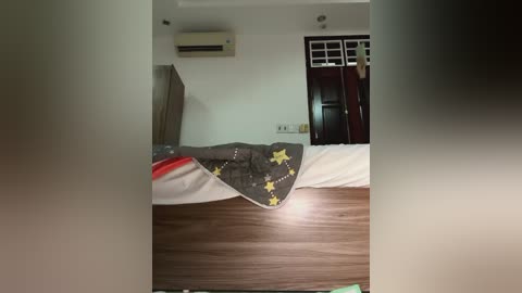 Media: Video of a small, modern bedroom with a wooden bed covered in a dark blanket with yellow stars, a white wall, an air conditioner, and a closed door with windows.