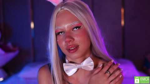 Media: Video of a young woman with long platinum blonde hair, wearing a white bunny ears headband, a bow tie, and makeup with pink eyeshadow, against a dimly lit purple background.