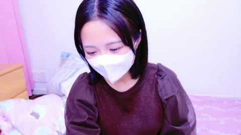 Media: Video of a young East Asian woman with straight black hair, wearing a black long-sleeve shirt and a white surgical mask, sitting on a pink-patterned bed, in a brightly lit room with pastel-colored walls.