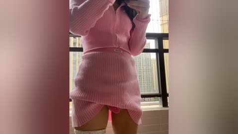 Media: Video of a light-skinned woman in a pink ribbed sweater dress, lifting the skirt to reveal pink underwear, standing in front of a window with city skyline.