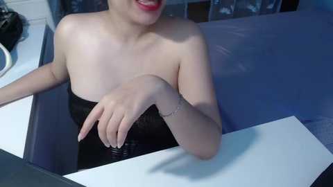 Media: Video of a smiling Asian woman with fair skin, wearing a strapless black dress, sitting in a bathtub, water splashing on the white countertop, with a blurred background.