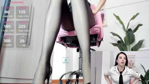 Media: Video of a woman in a pink chair, wearing black stockings and high heels, with a desk and plant in the background.