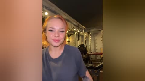 Media: Video of a young woman with light skin and shoulder-length red hair, wearing a gray T-shirt, standing in a dimly lit gym with mirrors, exercise machines, and fluorescent lights.