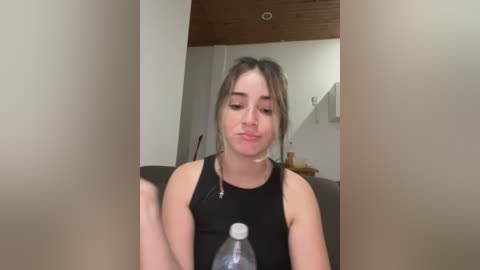 Media: Video of a young woman with light skin and shoulder-length blonde hair, wearing a black tank top, sitting in a dimly lit living room with beige walls and a wooden ceiling. She's drinking from a plastic water bottle.