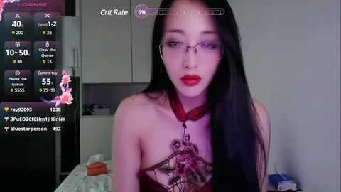 Media: Video of a pale-skinned woman with long black hair, red lipstick, and glasses, wearing a black lace top with a floral design, in a dimly lit room.