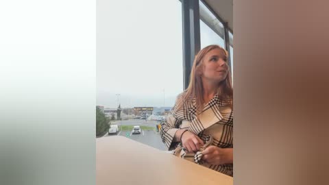 Media: Video of a blonde woman in a plaid coat, looking out of a large window at a cloudy sky and urban landscape, holding a clipboard.