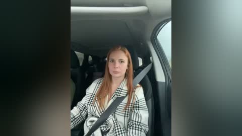 Media: Video of a young woman with long, straight red hair, wearing a black-and-white checkered coat, seated in a car, seatbelt on, looking forward. The interior is dimly lit, with gray and black car seats visible.