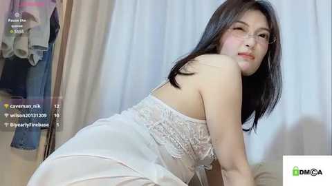 Media: Video of an East Asian woman with long black hair, wearing a white lace dress, leaning on a bed, indoors.