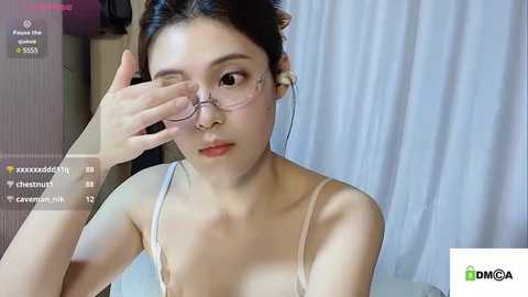 Media: A video of an East Asian woman with fair skin, wearing glasses, adjusting them. She has dark hair tied back and is in a white tank top. The background is a blurred, neutral-toned room.