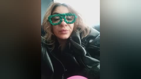 Media: Video of a woman with shoulder-length wavy hair, wearing green crocheted cat-eye glasses and a black leather jacket, sitting in a car.