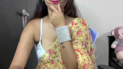 Media: Video of a young woman with medium skin tone, wearing a floral yellow top, exposing a light blue bra. She has a tattoo on her left wrist. Background features a door, stuffed animals, and a shelf.