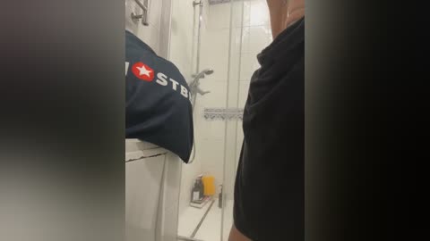 Media: Video of a person in a bathroom, wearing a black shirt, holding a blue towel with a red star logo. The background shows a white tiled wall, a sink, and a glass shower door.