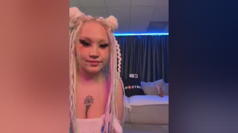 Media: Video of a young woman with light skin and platinum blonde dreadlocks, wearing a white top, smiling, in a dimly lit, modern living room with a couch and curtains.