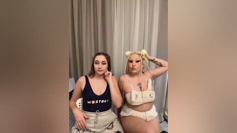 Media: Video of two young women with light skin and medium build, wearing casual outfits, seated against a beige curtain background.