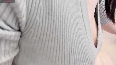 Media: Video of a close-up of a woman\u2019s ribbed, light gray sweater, highlighting the textured fabric. Partially visible is her dark hair and the top of her shoulder.